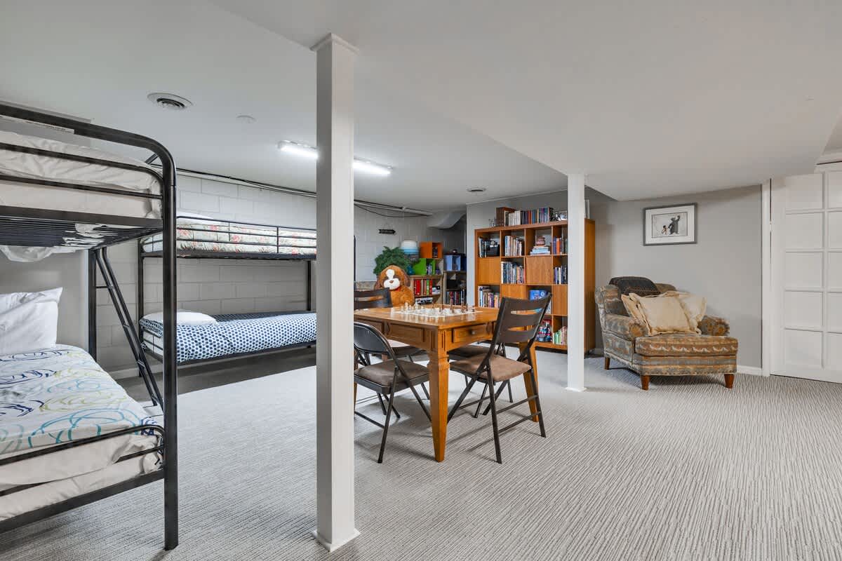 A wide shot of the basement game room, showcasing several metal bunk beds, a wooden game table with a chess set, and a cozy armchair next to a bookshelf. The room has light grey carpet and white support pillars.