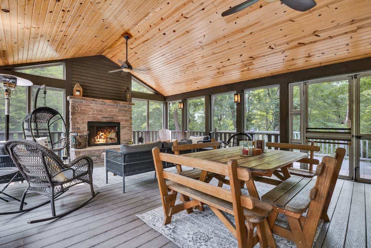 A spacious screened porch with a vaulted wood-paneled ceiling and dark gray walls, featuring a stone fireplace, a dining area with a rustic wooden table, and a lounge area with comfortable seating. Large windows offer expansive views of the surrounding trees.