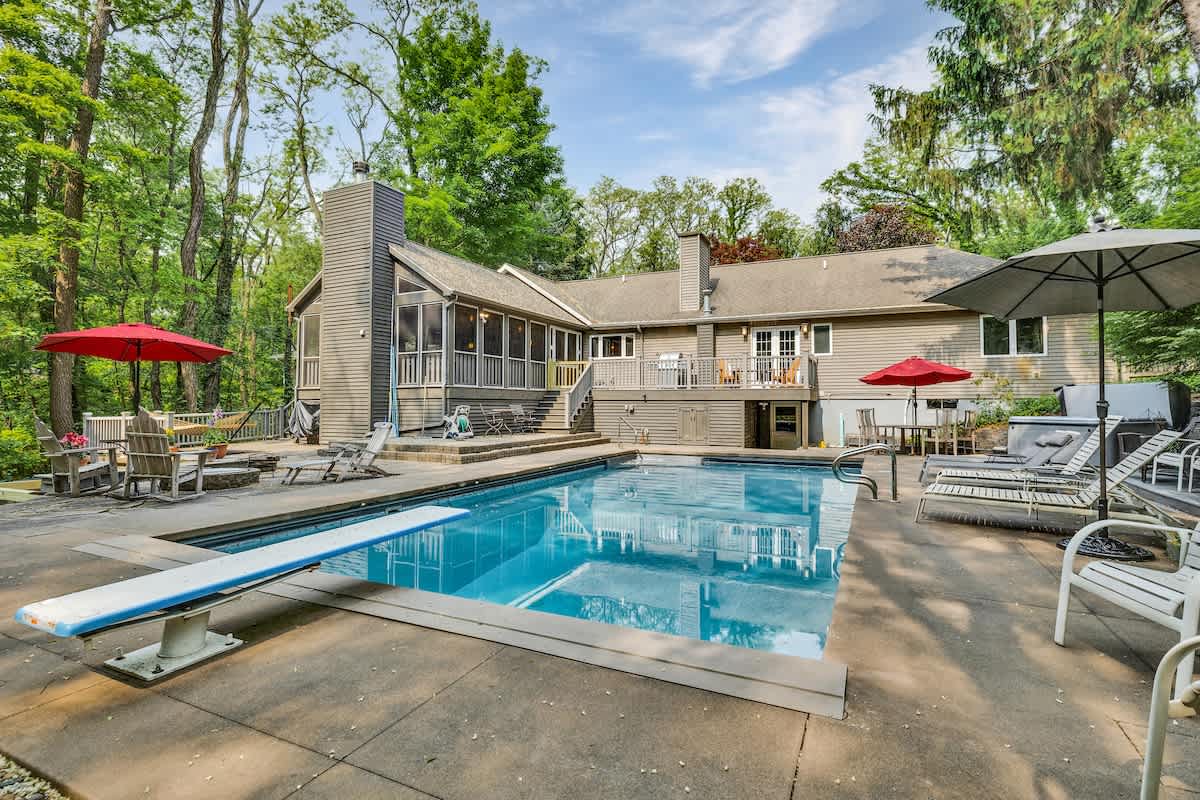 The back of the house features light gray siding and a large concrete patio surrounding a rectangular inground pool with a diving board. Red umbrellas provide shade over outdoor seating areas, all set against a lush green wooded backdrop.