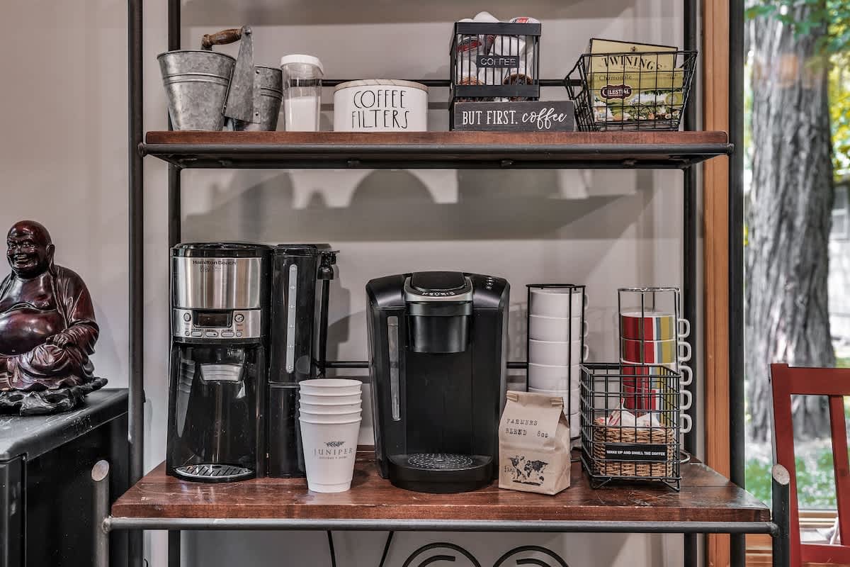 A dedicated coffee station features two coffee makers, a Keurig and a drip machine, along with various mugs, filters, and coffee supplies on a rustic metal and wood shelving unit. A Buddha statue sits to the left.