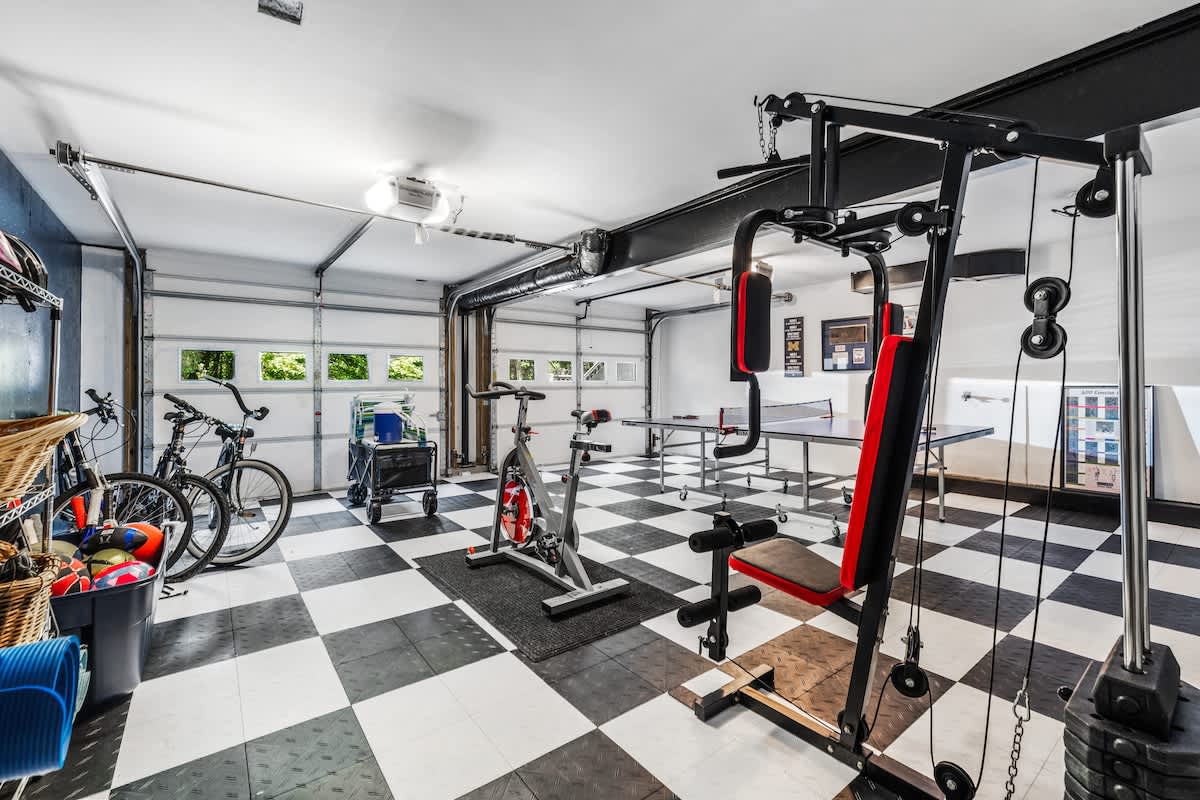 A spacious garage converted into a multi-functional gym and game room, featuring black and white checkered floor tiles, a multi-station gym machine, a stationary bike, and a ping-pong table. Garage doors are visible in the background.