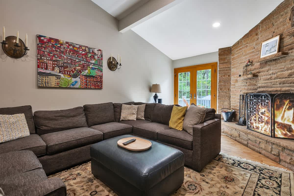 The living room is anchored by a floor-to-ceiling stone fireplace with a rustic mantel. A large dark gray sectional sofa and ottoman are arranged on an ornate area rug, with light gray walls and a vaulted ceiling.