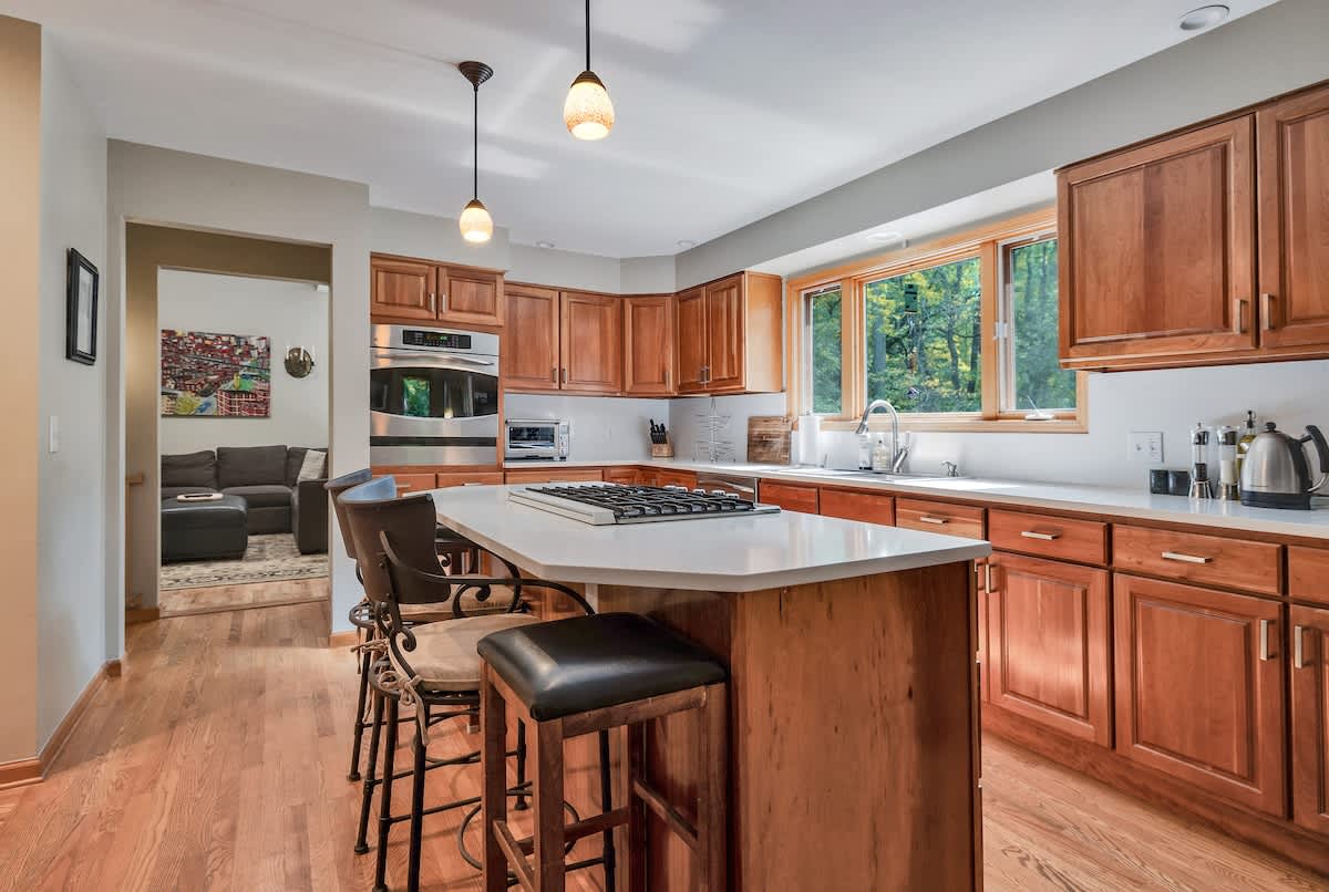 View of the kitchen showcasing light wood cabinetry, white countertops, and a central island with a gas range and bar seating. An open doorway leads to an adjacent living area, and hardwood floors extend throughout.