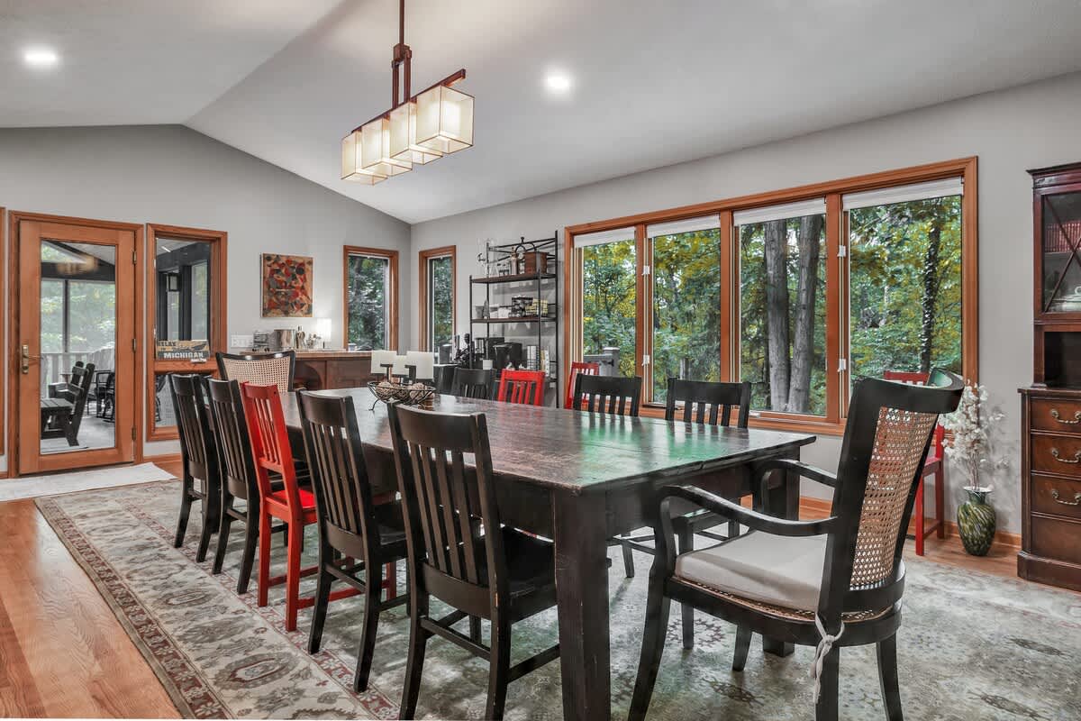 An expansive dining room features a long, dark wood dining table with an eclectic mix of chairs, including some with red accents. Large windows offer a view of the surrounding forest, and a modern rectangular chandelier hangs above.