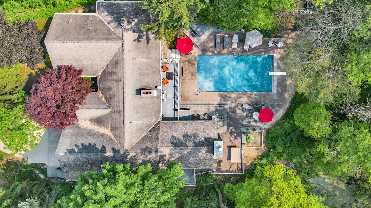 An aerial view showcases the large house with a gray shingled roof, surrounded by dense green trees. A rectangular inground pool is prominent in the backyard, along with multiple outdoor seating areas and red umbrellas.
