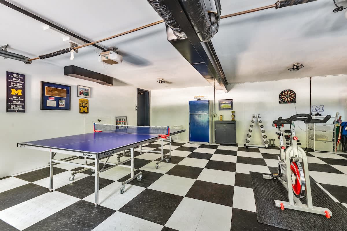 A spacious game room, likely a converted garage, features a black and white checkered floor, a ping-pong table, an exercise bike, and various gym equipment. University of Michigan memorabilia adorns the white walls.