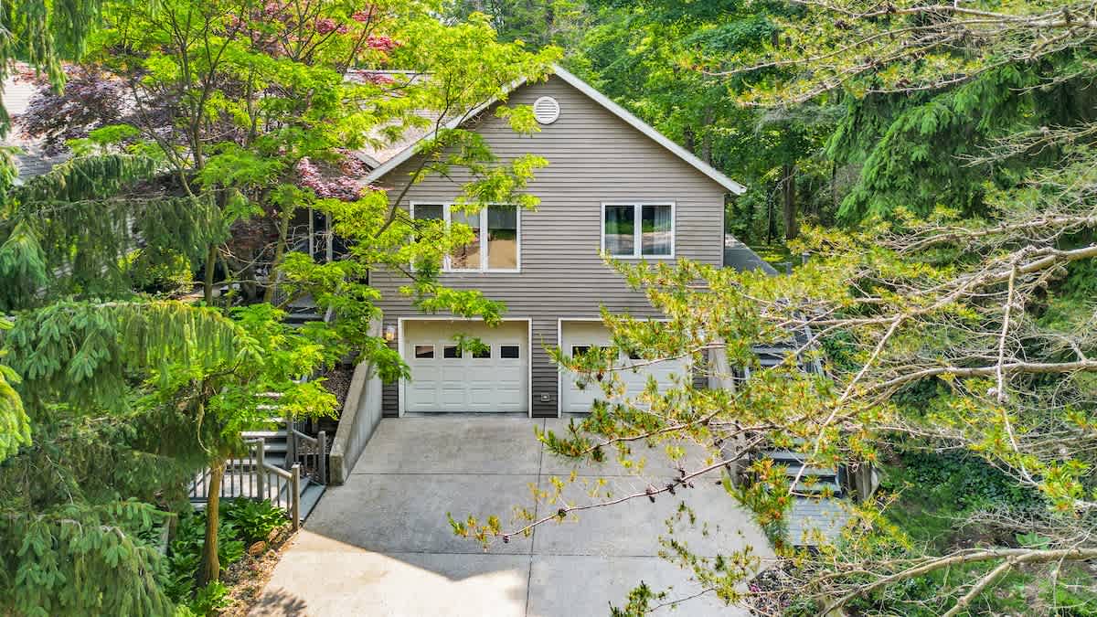 An elevated view of the multi-level house, showcasing two white garage doors and a concrete driveway, nestled among abundant green trees and foliage.