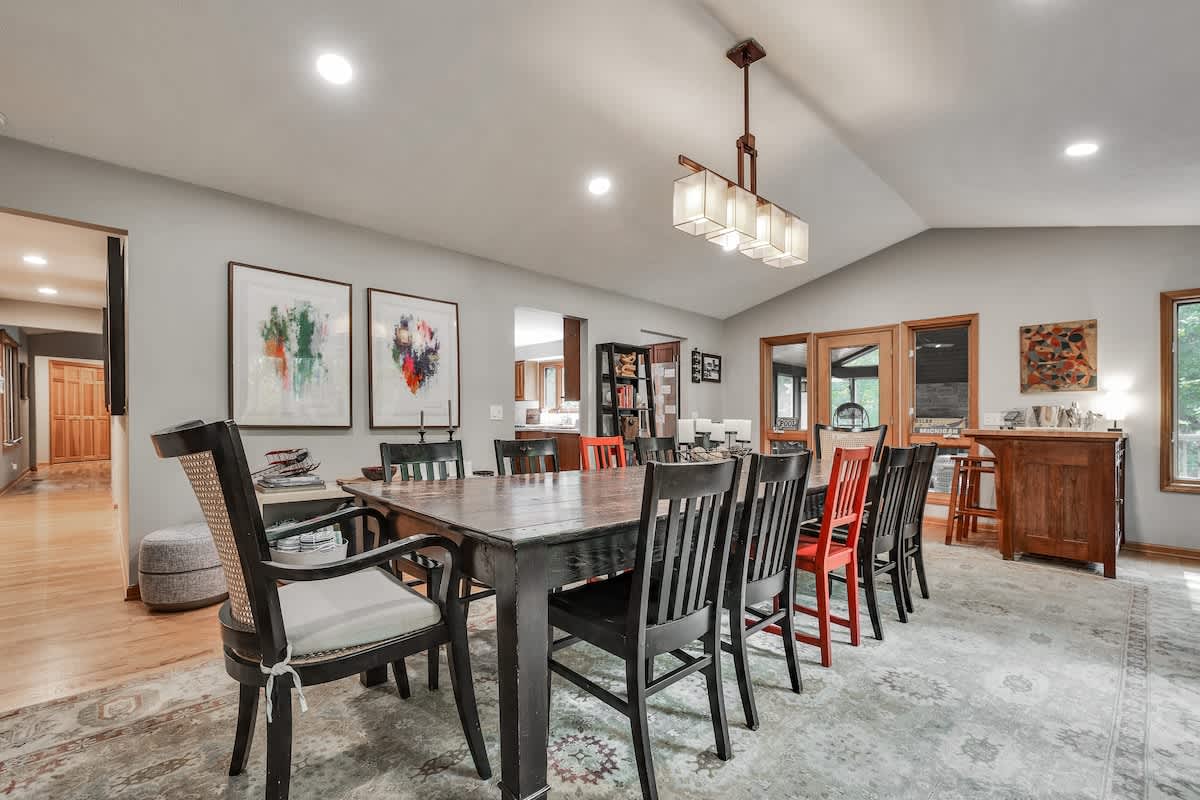 A spacious dining room features a large dark wood table surrounded by a mix of black and red chairs, set on a patterned area rug under a vaulted ceiling with recessed lighting and a modern chandelier. Two large abstract art pieces adorn the light gray wall.