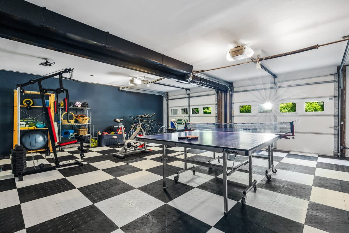 A wide shot of the garage game room, highlighting a blue ping-pong table in the center, surrounded by black and white checkered floor tiles. Bicycles and gym equipment are visible along the dark blue accent wall.