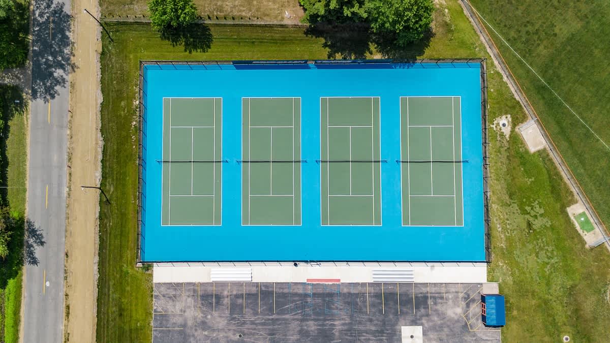 An aerial view of four vibrant tennis courts, featuring blue outer areas and green playing surfaces, surrounded by a fence, a parking lot, and a road.