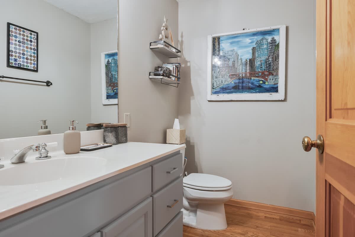 A modern bathroom with a light grey vanity, white countertop, and a large mirror. The walls are light grey, and the floor is hardwood. Artwork depicting a city scene hangs on the wall, adding a touch of urban flair.