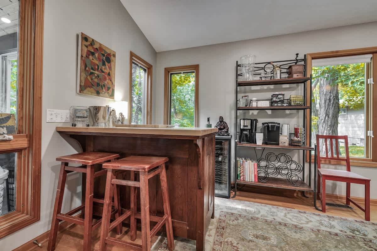 A cozy bar area with a wooden counter and two matching bar stools, featuring a wine cooler, coffee makers, and a shelving unit displaying various glassware and decor. Large windows offer views of the outdoors.