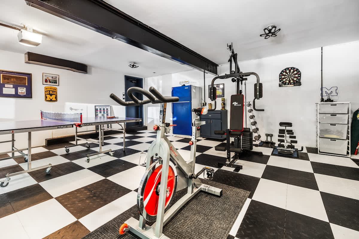 A comprehensive view of the garage gym and game room, featuring a stationary bike, a multi-station gym, free weights, and a ping-pong table. A vintage blue refrigerator and a dartboard add character to the space with checkered floor tiles.