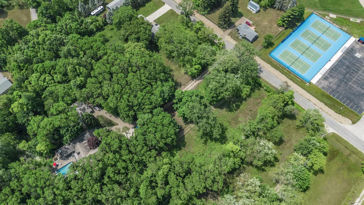 Overhead aerial view highlighting the secluded house and pool within a vast wooded area, with bright blue tennis courts and other residential structures visible in the upper right background.