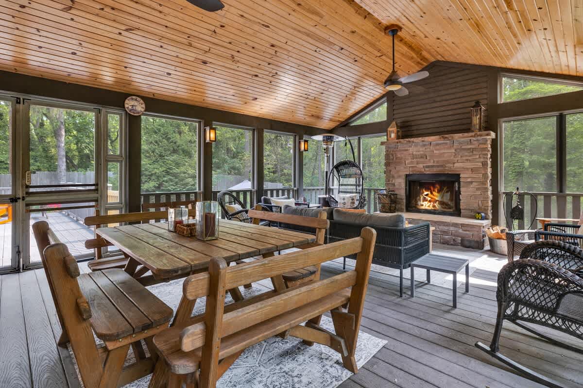 A spacious screened-in porch with a vaulted wood-paneled ceiling features a rustic wooden dining table and benches, a cozy seating area with a stone fireplace, and large windows overlooking the surrounding trees.