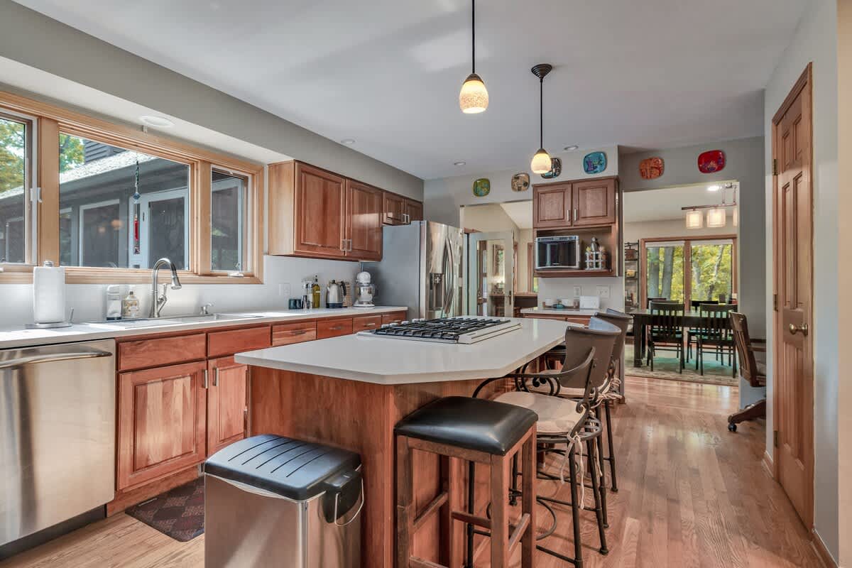 A bright kitchen with light wood cabinetry, white countertops, and stainless steel appliances, including a large island with a gas cooktop and seating for three. Pendant lights hang above the island, and a large window overlooks greenery.