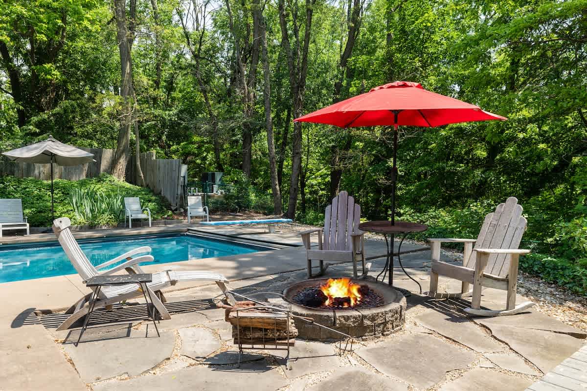 Outdoor patio with a swimming pool, a lit fire pit surrounded by Adirondack chairs, and a red umbrella, all set amidst lush green trees.