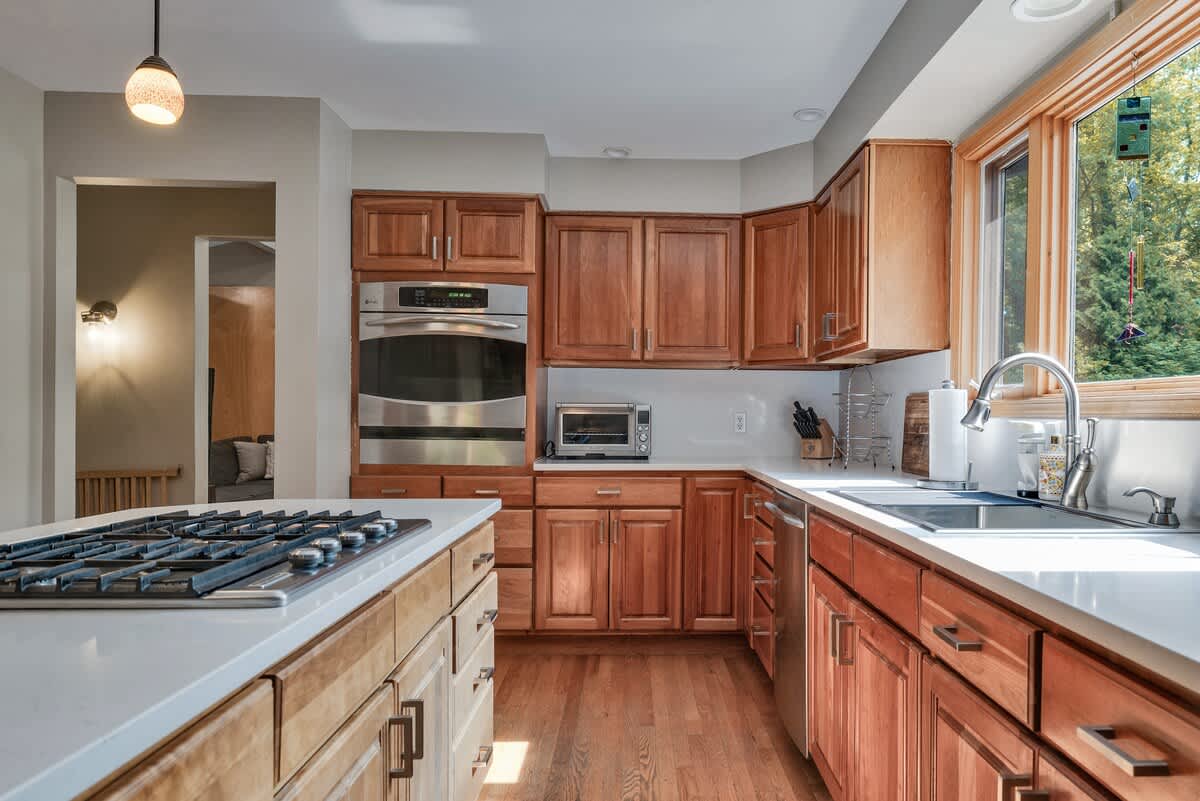 Close-up of the kitchen featuring light wood cabinetry, white countertops, and stainless steel appliances including a built-in oven, microwave, and dishwasher. A large window above the sink provides natural light.