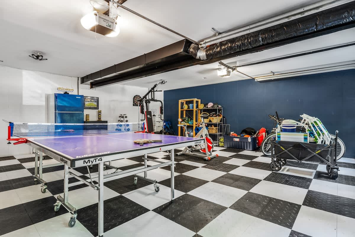 A spacious game room featuring a purple ping pong table, black and white checkered floor tiles, and various gym equipment and storage shelves against a dark blue accent wall.