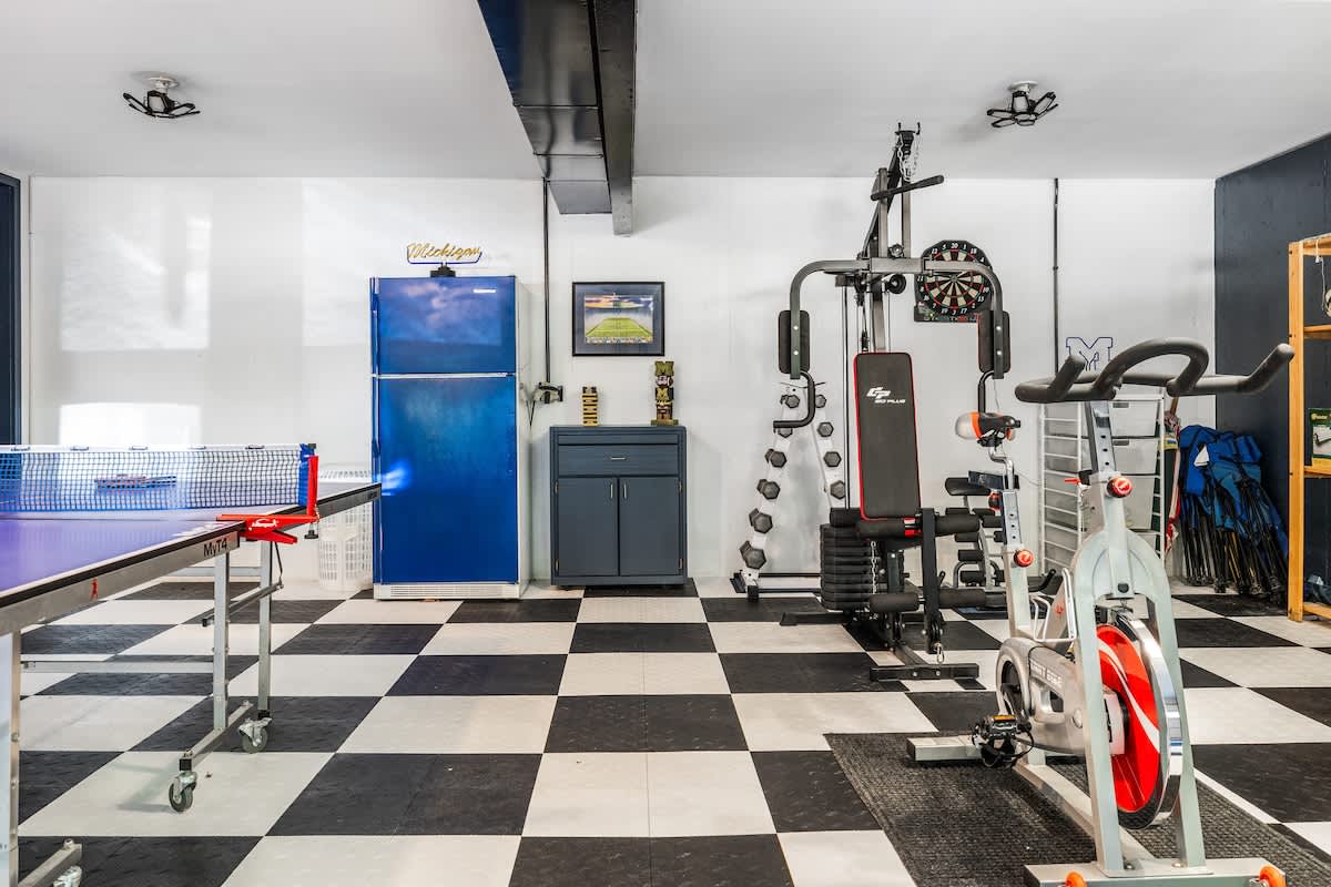 A detailed shot of the garage gym area, showcasing a multi-station gym machine, free weights, a stationary bike, and a vintage blue refrigerator. The black and white checkered floor tiles extend throughout the space.