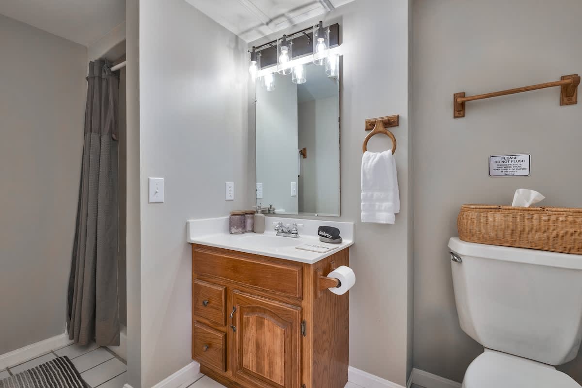 A compact bathroom featuring a wooden vanity with a white countertop and sink, a large mirror with a modern light fixture, a toilet, and a shower with a grey curtain.