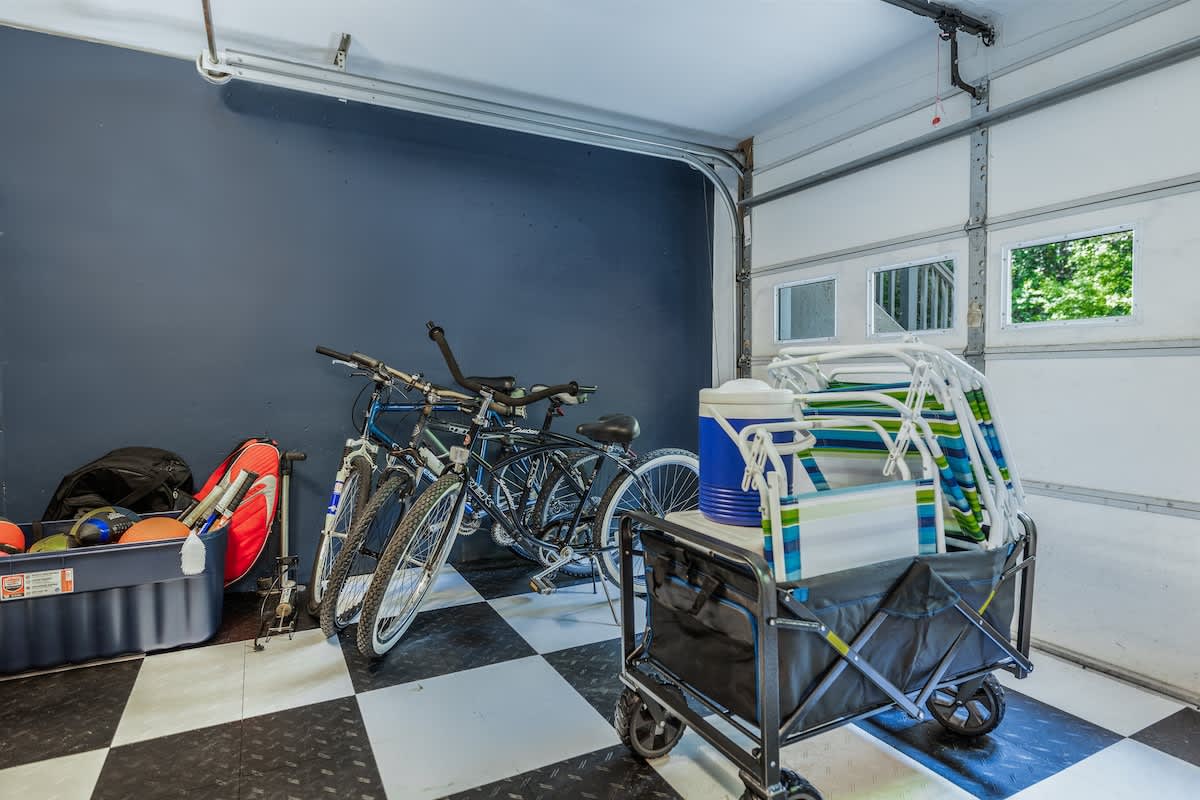 A section of the garage dedicated to storage and outdoor gear, featuring several bicycles, a utility cart filled with beach chairs and a cooler, and various sports equipment. The floor has black and white checkered tiles.