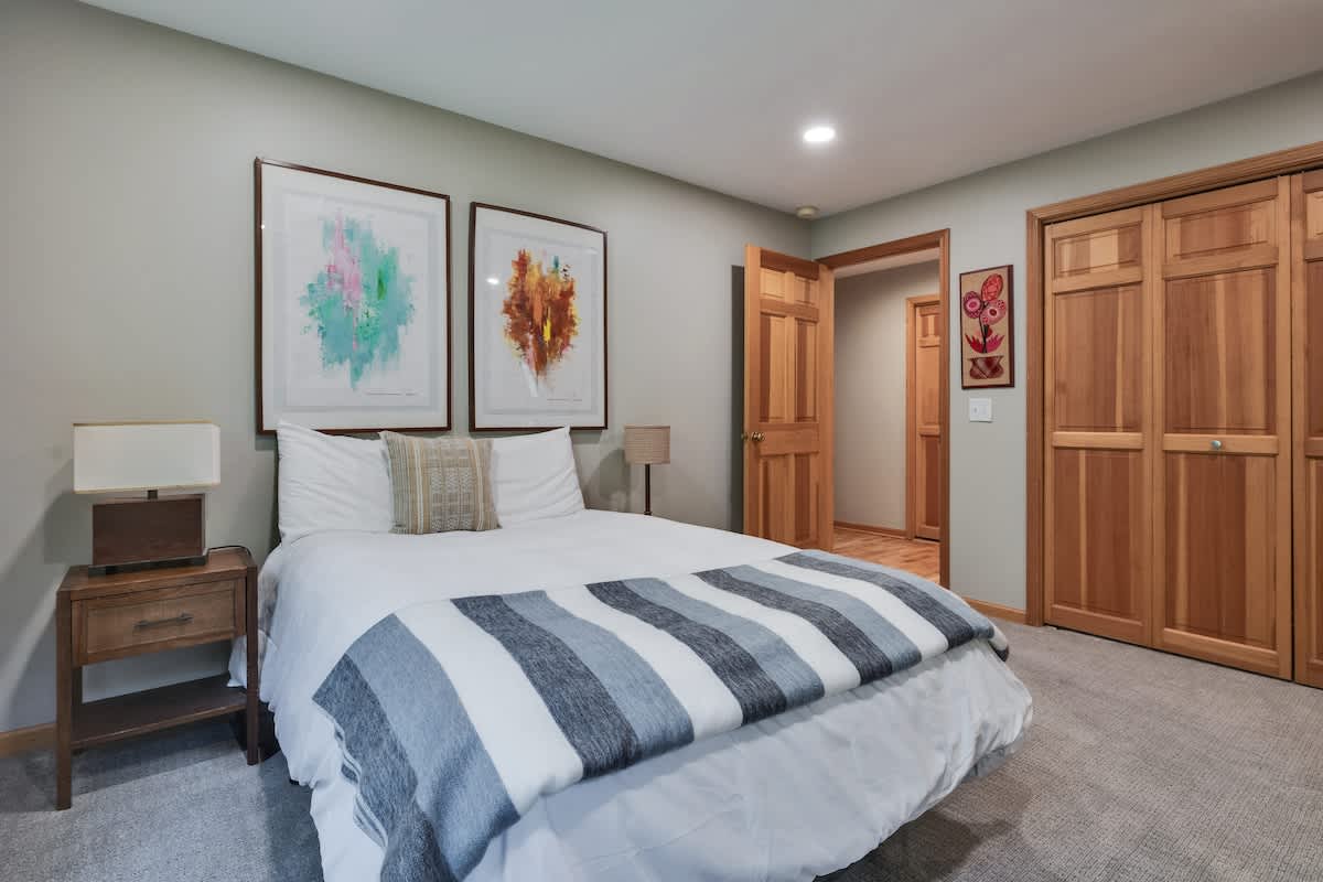 A cozy bedroom featuring a queen-sized bed with white bedding and a blue and grey striped throw, flanked by a wooden nightstand with a lamp. Two large abstract art pieces hang above the bed, complemented by light grey walls and carpet.