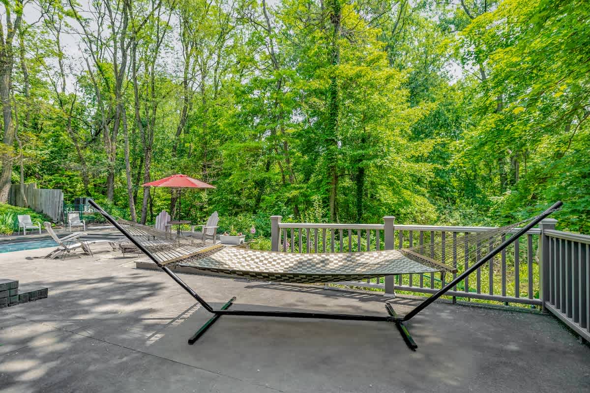 A comfortable hammock on a concrete patio, overlooking a wooden deck with railings, a swimming pool in the distance, and surrounded by a dense, vibrant green forest.