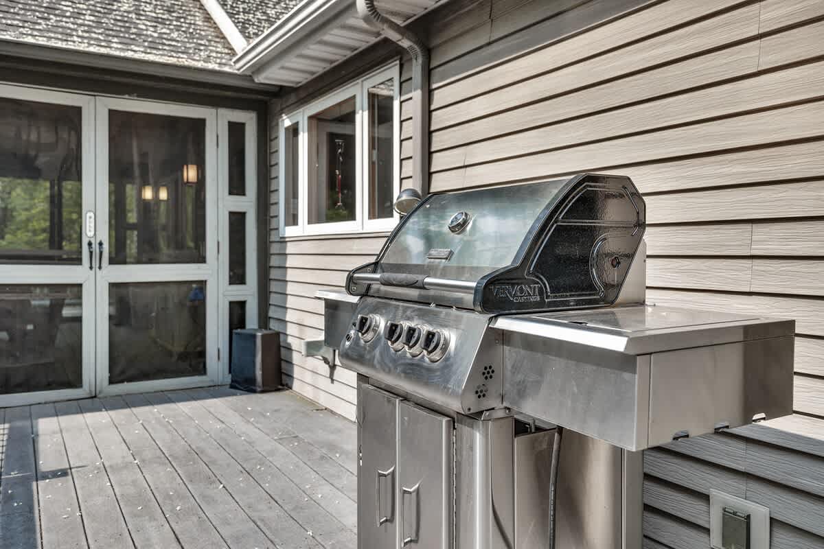 A stainless steel BBQ grill sits on a gray wooden deck next to the house, which features light gray siding and large screened doors leading to an interior space. Lush greenery is visible in the background.
