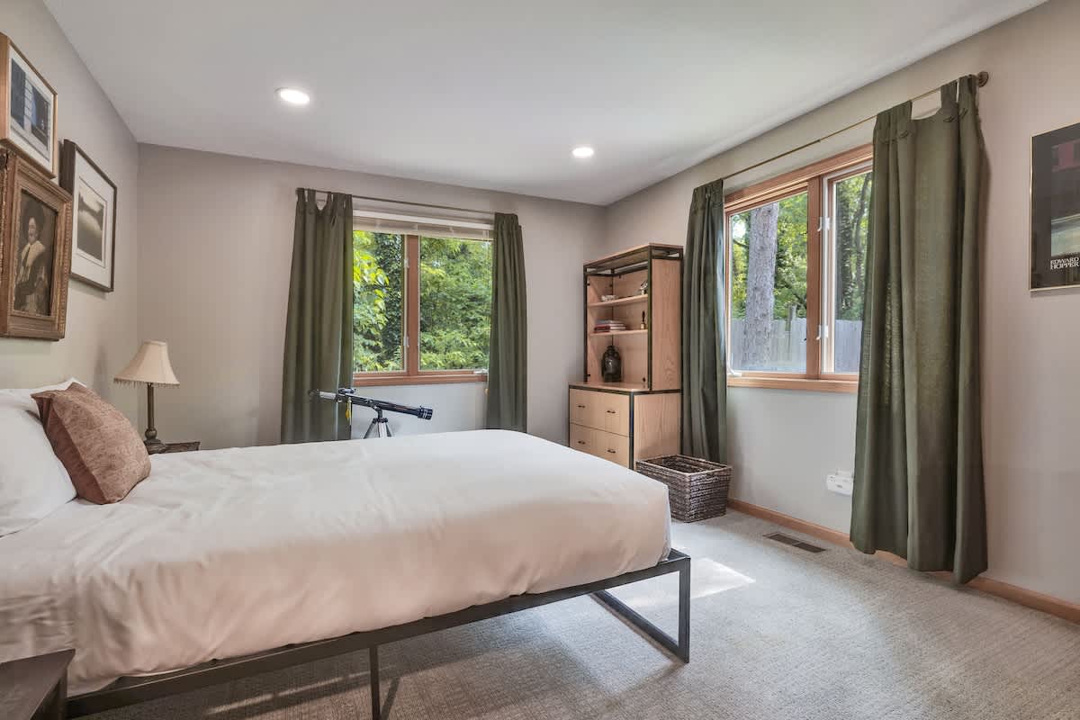 Bedroom featuring a queen-sized bed, two windows with dark green curtains, a wooden shelving unit with drawers, and a telescope near the window.