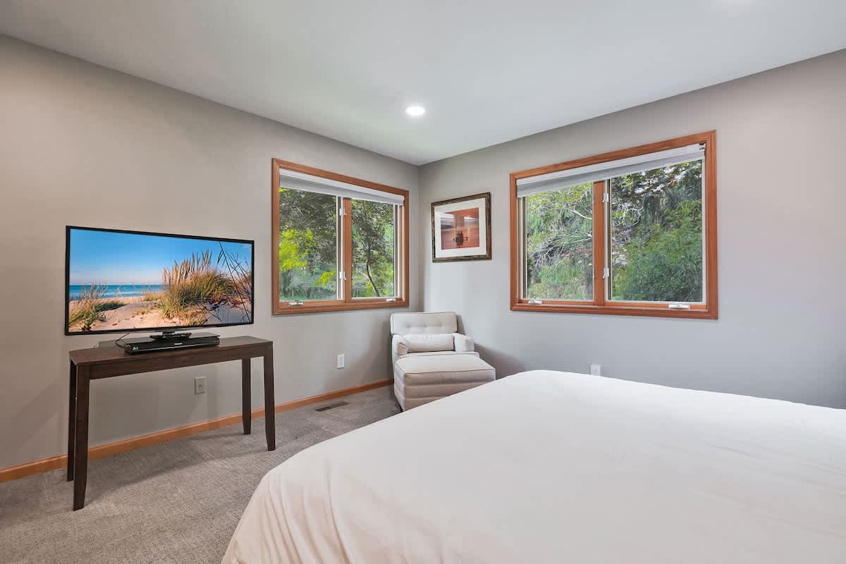 View of a bedroom with a king-sized bed, a large flat-screen TV on a console, a chaise lounge, and two windows offering views of lush trees.