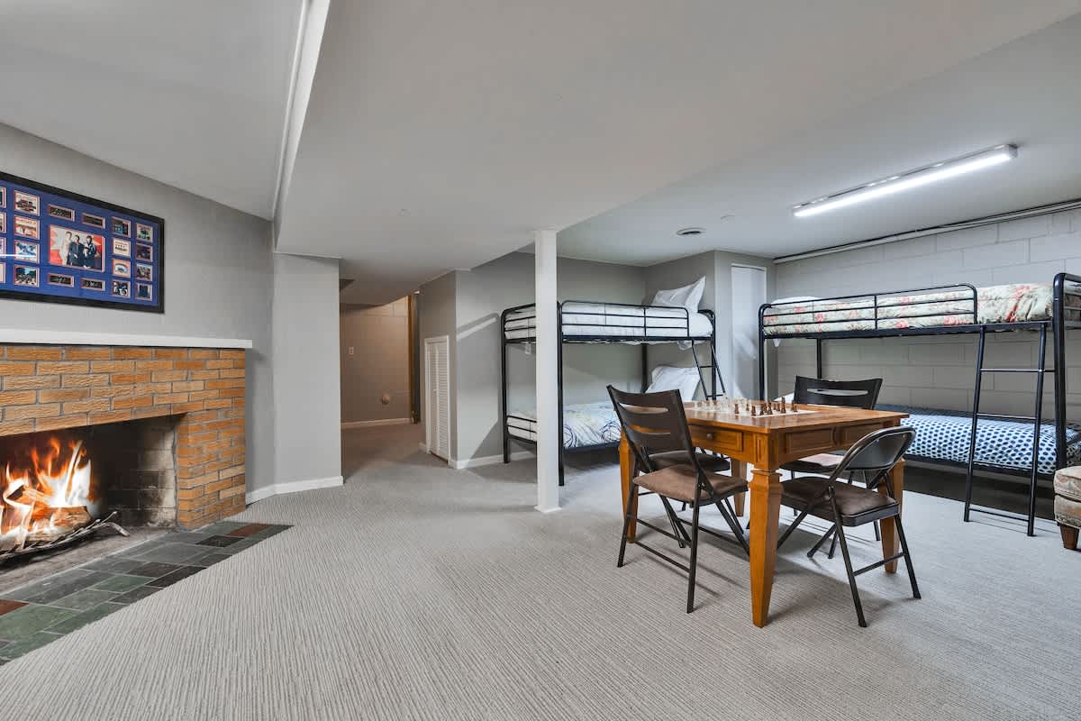 A spacious basement game room with light grey carpet, featuring a brick fireplace with a roaring fire, multiple metal bunk beds, and a wooden game table with chairs. The walls are light grey, and exposed ceiling beams add a rustic touch.