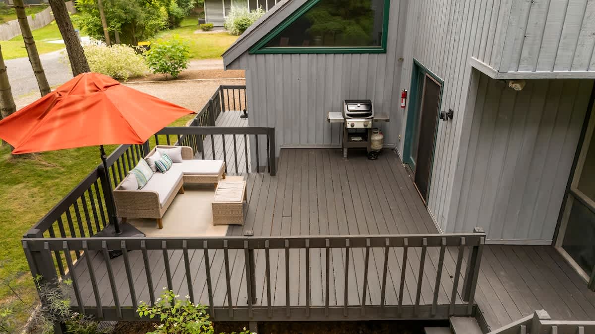 An elevated outdoor deck with dark grey wooden planks and railings, featuring a wicker sectional sofa with cushions under an orange umbrella, and a stainless steel BBQ grill. The house exterior is grey siding.