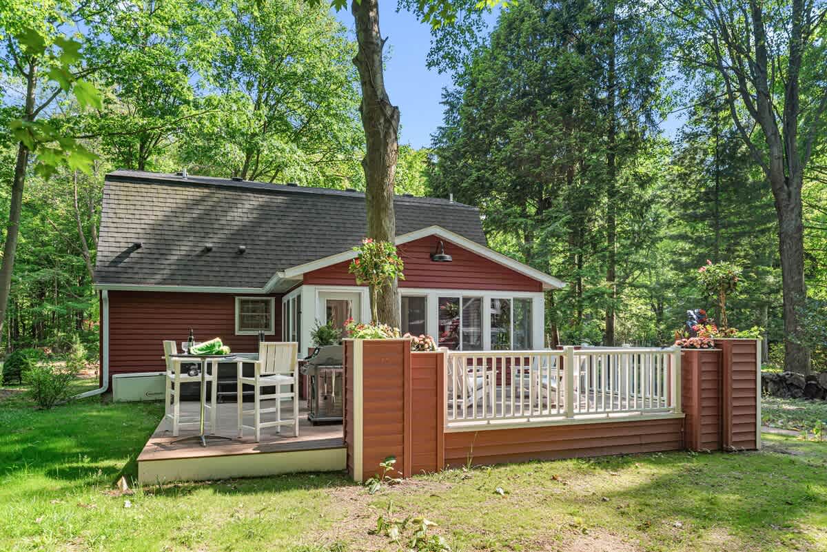 The back of a red-sided house with a wooden deck, featuring a hot tub and a high-top dining table with chairs, all set against a backdrop of lush green trees.