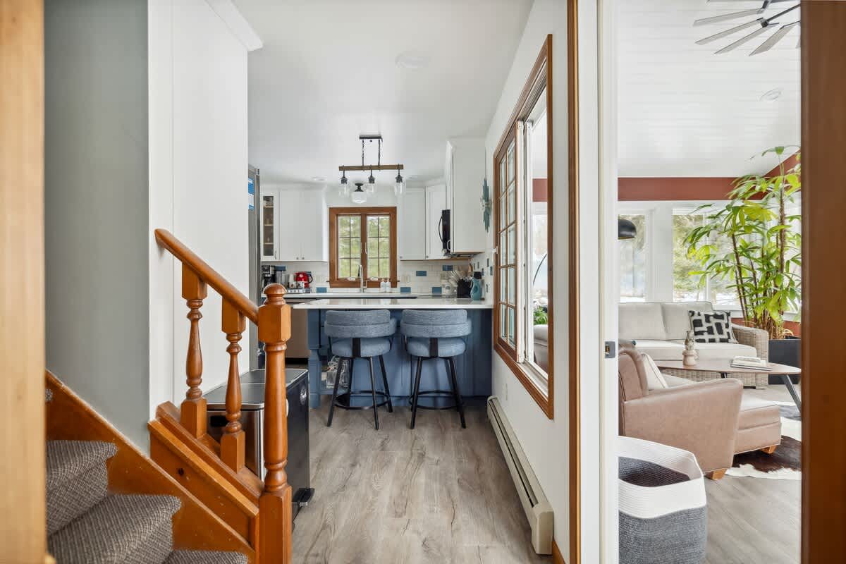 A carpeted staircase with a wooden railing leads up to an open-concept kitchen featuring blue and white cabinetry and a breakfast bar with stools. A separate living area with a light-colored sofa is visible through a doorway.