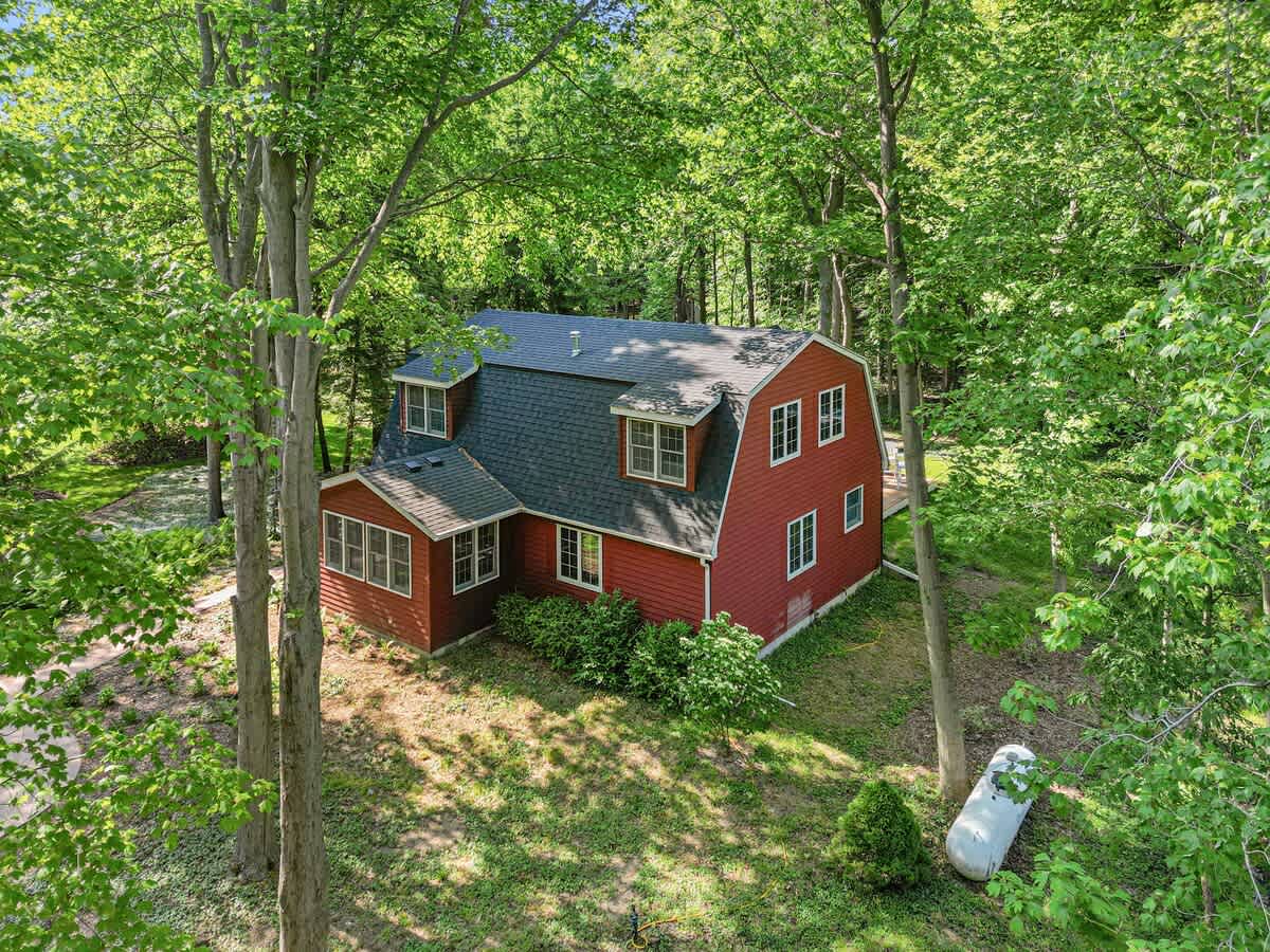 An aerial view of a red-sided house with a dark roof, nestled among dense green trees, showcasing the property's secluded, wooded setting. A propane tank is visible in the foreground.