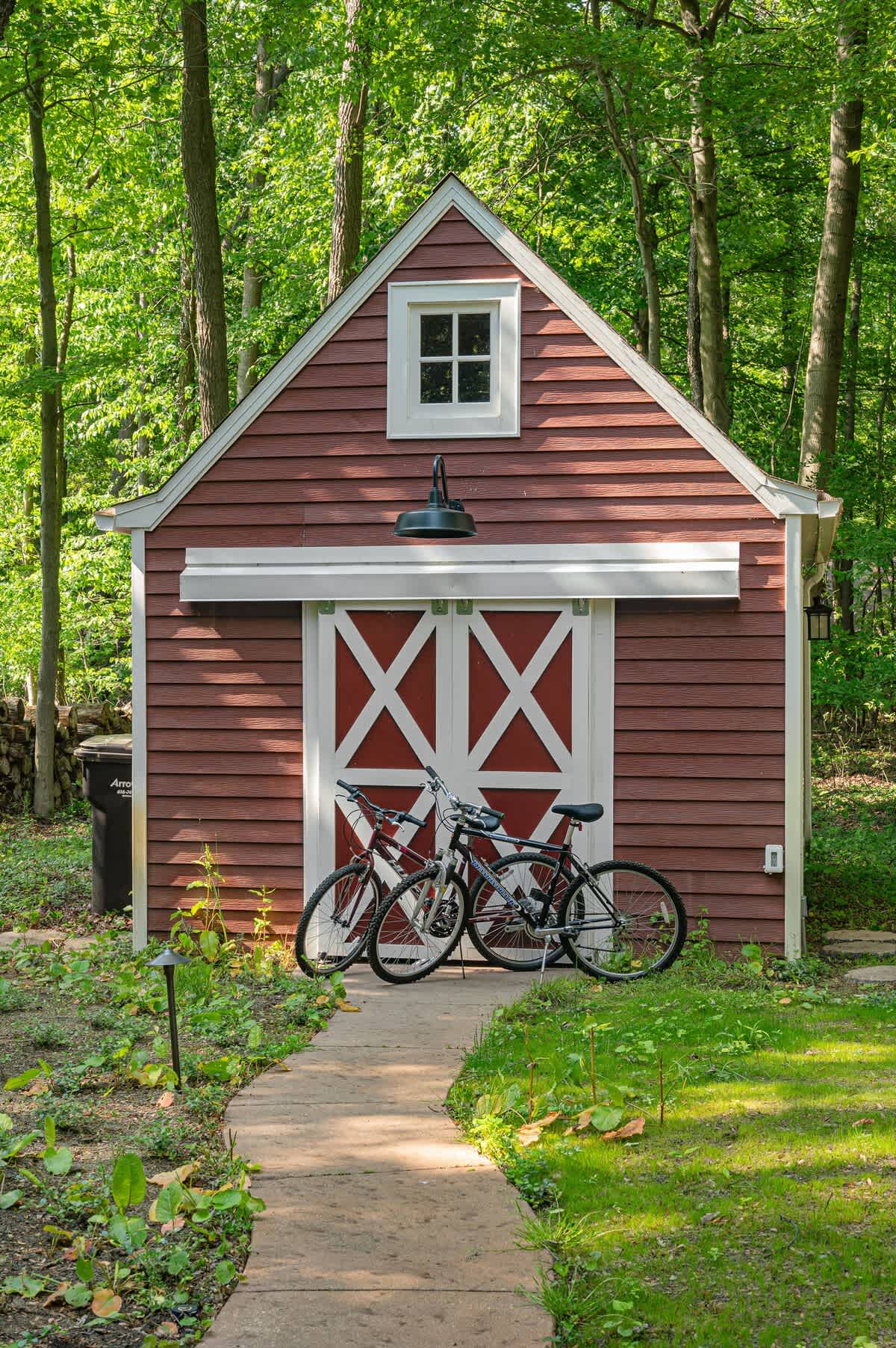 A charming red shed with white trim and classic barn-style doors, topped with a small window, is nestled in a lush green yard surrounded by mature trees. Two bicycles are casually parked in front, suggesting opportunities for outdoor recreation.