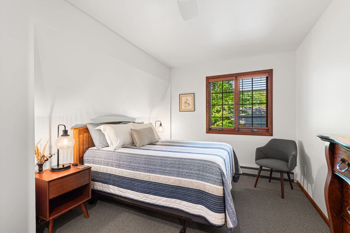 A cozy bedroom features a queen-sized bed with a wooden headboard and a blue and white striped comforter, flanked by a wooden nightstand with a modern lamp. A wooden-framed window with blinds overlooks greenery, and a gray armchair sits in the corner.