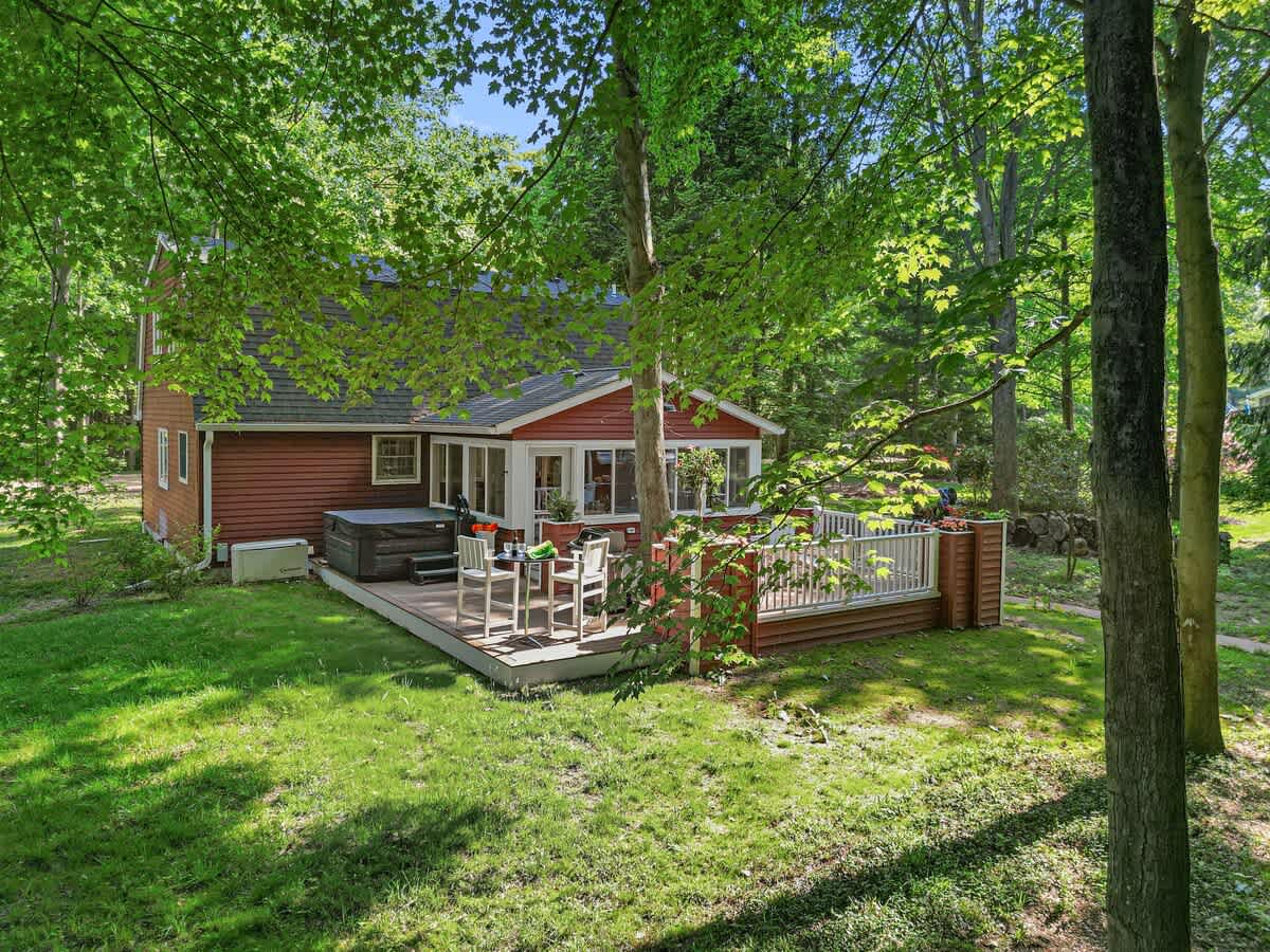An elevated view of the red-sided house with a large wooden deck, featuring a hot tub and outdoor dining area, surrounded by a vibrant green lawn and mature trees.