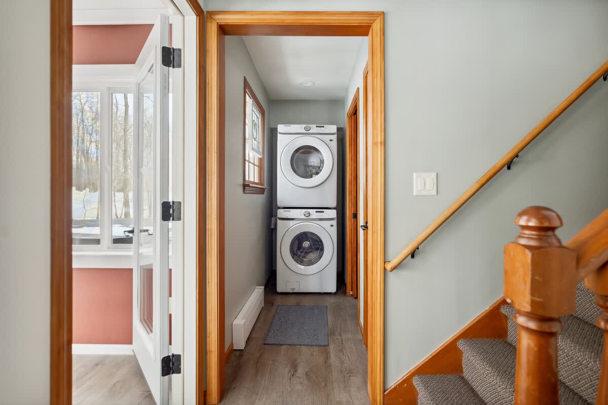 Hallway laundry area with light blue walls and wood-look flooring, featuring a stacked white washer and dryer unit. Wooden trim frames the doorways and a small window, with a staircase visible to the right.