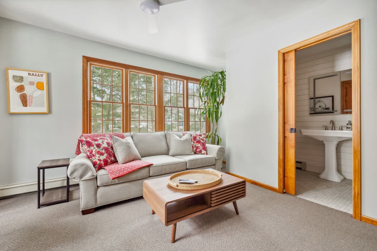 The living room features a light gray sofa with red floral pillows and a wooden coffee table, set against light gray walls and carpet. A doorway with wooden trim leads to a bathroom with a white pedestal sink.