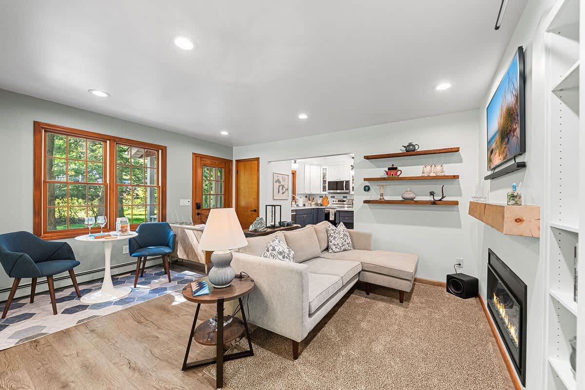 A spacious living room features a light-colored sectional sofa on carpet, facing a fireplace and wall-mounted TV. To the left, a bright dining nook with a white table and blue chairs is visible on hardwood floors, near large wood-framed windows.