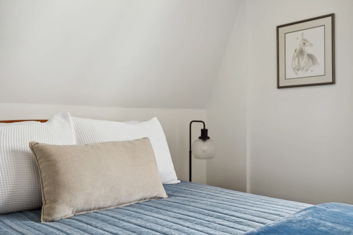 A close-up view of the bed in bedroom-2, featuring a white waffle-knit duvet, a blue quilted comforter, and a beige lumbar pillow. A modern black table lamp and framed artwork are visible on the white wall.