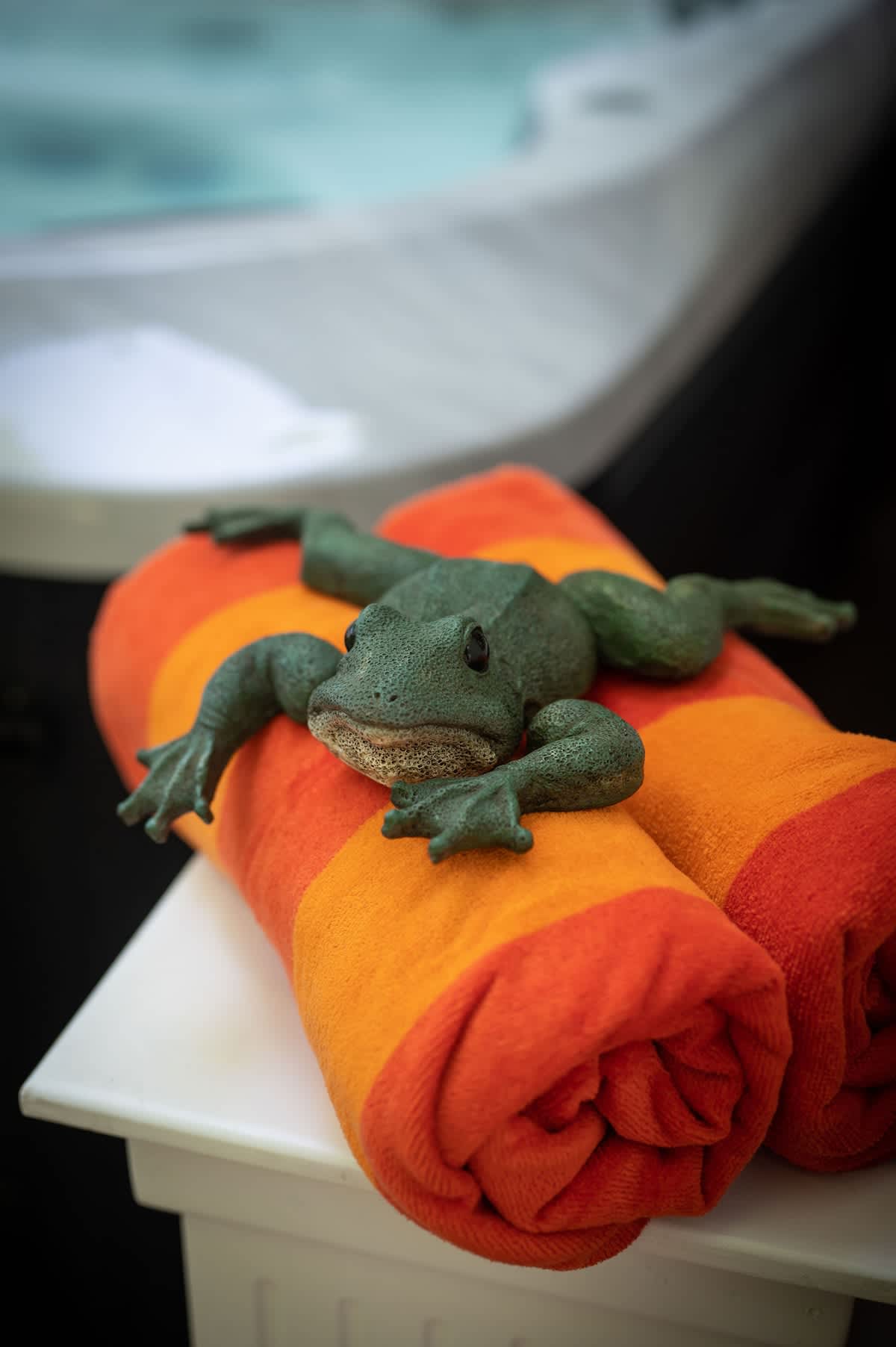 A close-up of a green frog statue resting on two rolled-up orange and red striped towels, with the blurred background of a hot tub.