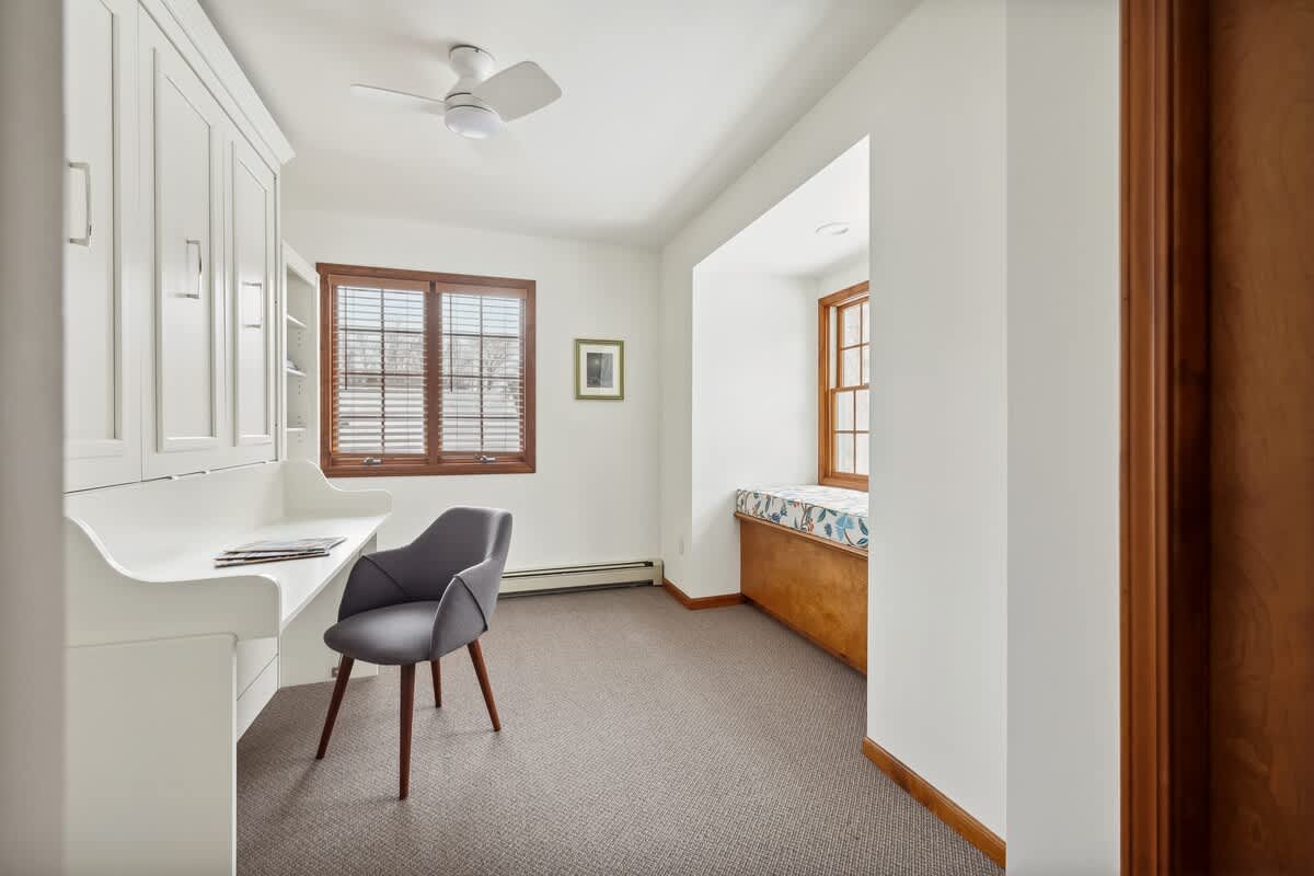 A bright home office featuring white built-in cabinetry with a desk, a gray upholstered chair, and a cozy window seat with a patterned cushion. Wooden blinds cover the windows, and the room has light gray carpet.