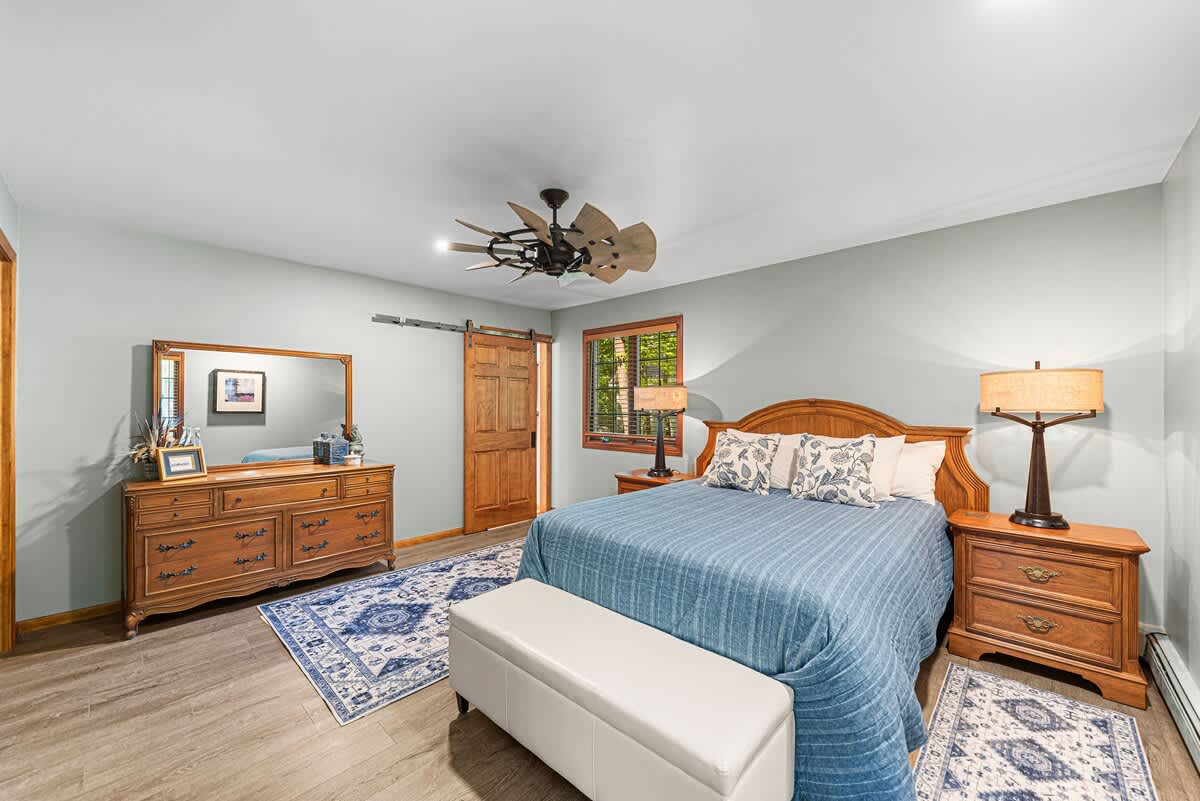Spacious bedroom featuring light blue walls, wood-look flooring, and a wooden bed frame with blue bedding. Includes a large wooden dresser with a mirror, two nightstands with lamps, and a unique ceiling fan.