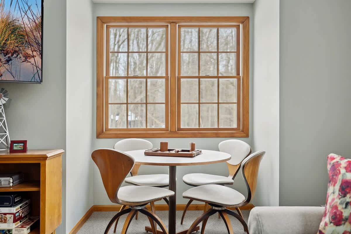 A cozy dining nook features a round white table with a wooden base, surrounded by four modern chairs with white seats and curved wooden backs. A large wooden-framed window with multiple panes overlooks a wooded outdoor scene.