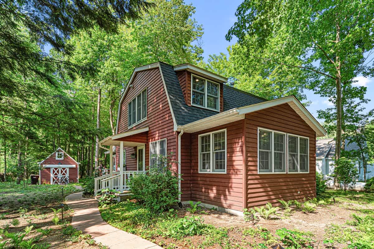 The inviting front exterior of a charming red house with white trim and a dark roof is surrounded by lush green trees and well-maintained landscaping. A concrete pathway leads to the welcoming front porch, with a matching red shed visible in the background.