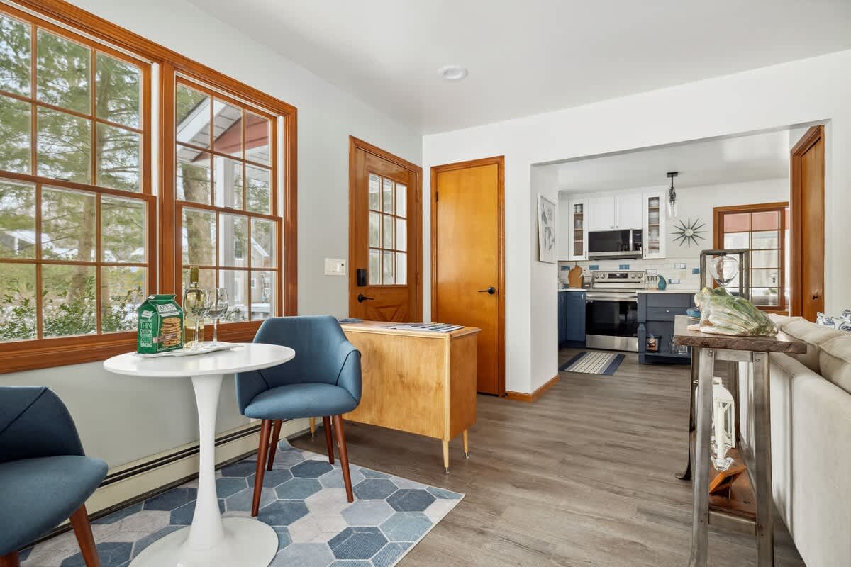 A bright dining nook features a white round table with two blue upholstered chairs on a blue and white geometric rug, set against large wood-framed windows. Beyond, a modern kitchen with blue and white cabinetry is visible.