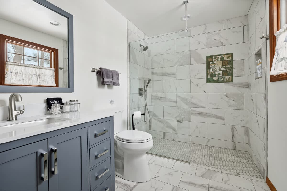 Modern bathroom featuring a grey vanity with a white countertop, a toilet, and a spacious walk-in shower with white marble-look subway tiles and a glass enclosure. The floor has matching marble-look tiles.